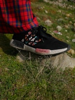 A close-up view of a foot wearing a black shoe with floral designs and standing on a rock. The shoe has a pink section with text on the side. The person is wearing red plaid pants. The ground is grassy with some rocks in the background.