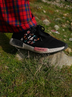 A close-up view of a foot wearing a black shoe with floral designs and standing on a rock. The shoe has a pink section with text on the side. The person is wearing red plaid pants. The ground is grassy with some rocks in the background.