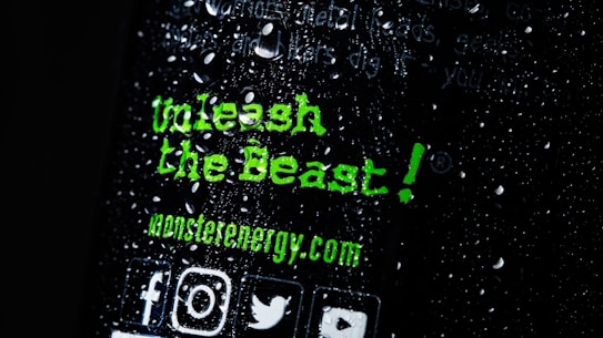 There is text written in a bold, green font that reads 'Unleash the Beast!' along with the website 'monsterenergy.com'. The background is dark with water droplets scattered across the surface. Several social media icons are visible at the bottom.