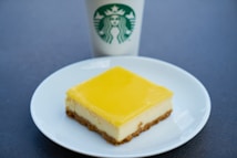 A square piece of lemon cheesecake on a white plate, featuring a creamy middle layer and crumbly base with a glossy yellow top layer. Behind the plate is a Starbucks cup with a green logo.