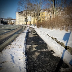 A snowy urban landscape featuring a partially snow-covered road alongside a cleared sidewalk. Tall, leafless trees line the path, and a row of classic European-style buildings is visible in the background. A road sign indicates a right turn ahead at 150 meters.