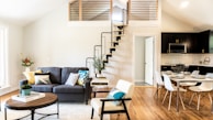 A modern living room and kitchen area features a cozy gray sofa with colorful cushions, a wooden coffee table, and an armchair with a cushion. A plant and some decorative items are arranged on the table. The space is enhanced by wooden flooring and a loft with a unique spiral staircase. The kitchen area includes a dining table set with dishes and chairs. The walls are painted in neutral tones, and natural light streams in through large windows.
