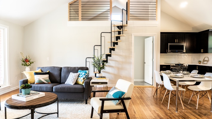 A modern living room and kitchen area features a cozy gray sofa with colorful cushions, a wooden coffee table, and an armchair with a cushion. A plant and some decorative items are arranged on the table. The space is enhanced by wooden flooring and a loft with a unique spiral staircase. The kitchen area includes a dining table set with dishes and chairs. The walls are painted in neutral tones, and natural light streams in through large windows.
