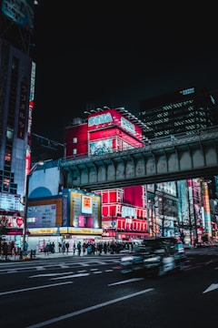 Neon-lit urban street scene with a prominent red and blue SEGA building, bustling pedestrians, and light trails from passing vehicles. Elevated railway tracks cross overhead, and surrounding buildings display brightly colored advertisements.