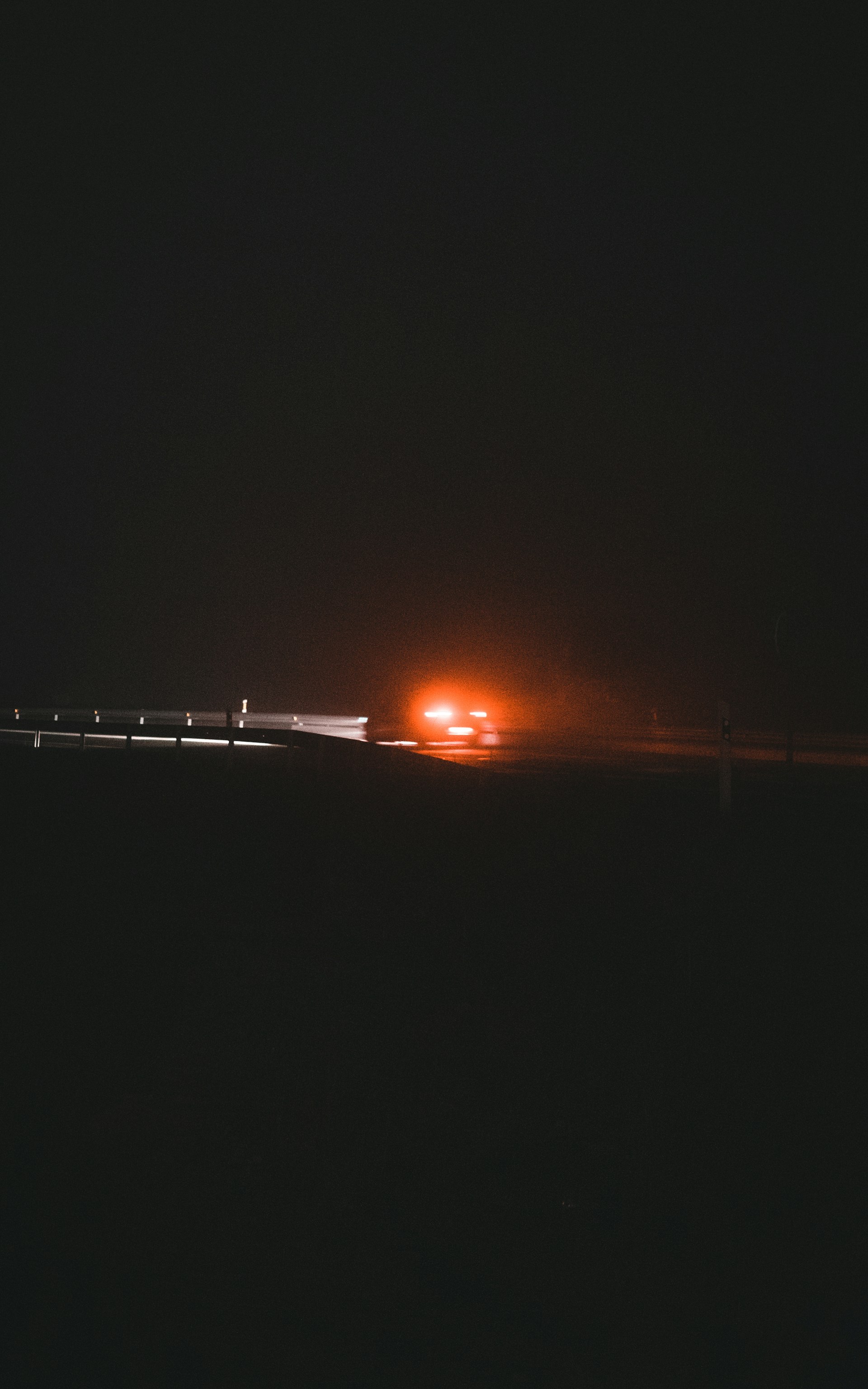 A car travels along a dark road at night, illuminated by the faint glow of its red brake lights. The scene is minimally lit, creating a mysterious and somewhat eerie atmosphere.