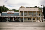 A quaint, two-story building with a traditional architectural style. The left side features a store named Mason's Healthcare, offering medicines, cosmetics, and gifts, while the right side is a place called Lantern Court Motels. Both sections of the building are adorned with hanging flower baskets and there is a backdrop of lush greenery with a partly cloudy sky.