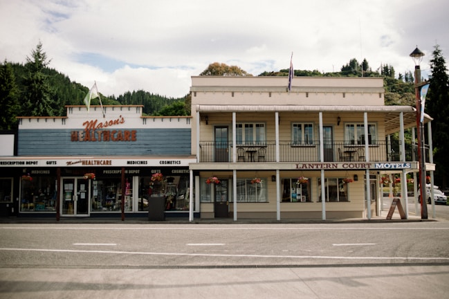 A quaint, two-story building with a traditional architectural style. The left side features a store named Mason's Healthcare, offering medicines, cosmetics, and gifts, while the right side is a place called Lantern Court Motels. Both sections of the building are adorned with hanging flower baskets and there is a backdrop of lush greenery with a partly cloudy sky.