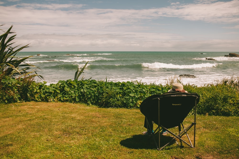 uomo seduto sulla sedia pieghevole nera sul campo di erba verde vicino al mare durante il giorno