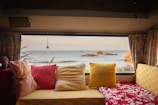 The image depicts the interior view of a cozy camper or RV, furnished with colorful cushions and pillows in shades of red, yellow, and beige. Through the large window, there is a scenic view of the ocean with a few visible rocks near the shoreline. Curtains frame the window, contributing to a relaxed and homey ambiance.
