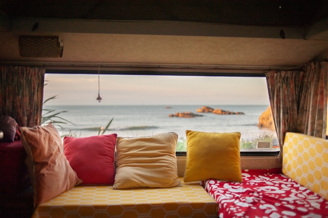 The image depicts the interior view of a cozy camper or RV, furnished with colorful cushions and pillows in shades of red, yellow, and beige. Through the large window, there is a scenic view of the ocean with a few visible rocks near the shoreline. Curtains frame the window, contributing to a relaxed and homey ambiance.