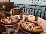 A seaside setting with two pizzas on a table. There are two chairs and two beer bottles, one open and one closed. Two glasses accompany the beverage, and a candle is placed in the center of the table. The ocean view in the background creates a relaxed atmosphere.