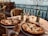 A seaside setting with two pizzas on a table. There are two chairs and two beer bottles, one open and one closed. Two glasses accompany the beverage, and a candle is placed in the center of the table. The ocean view in the background creates a relaxed atmosphere.