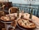 A seaside setting with two pizzas on a table. There are two chairs and two beer bottles, one open and one closed. Two glasses accompany the beverage, and a candle is placed in the center of the table. The ocean view in the background creates a relaxed atmosphere.