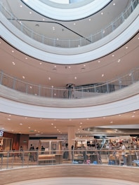 A multi-level shopping mall interior with open walkways and a circular atrium design. The scene includes several shoppers walking and visiting different stores, with bright ceiling lights illuminating the area.