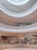 A multi-level shopping mall interior with open walkways and a circular atrium design. The scene includes several shoppers walking and visiting different stores, with bright ceiling lights illuminating the area.