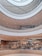 A multi-level shopping mall interior with open walkways and a circular atrium design. The scene includes several shoppers walking and visiting different stores, with bright ceiling lights illuminating the area.