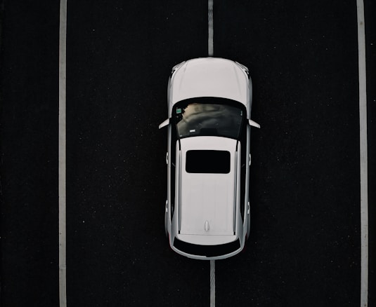 Aerial view of a white car parked in a space outlined by parallel white lines on a dark asphalt surface.