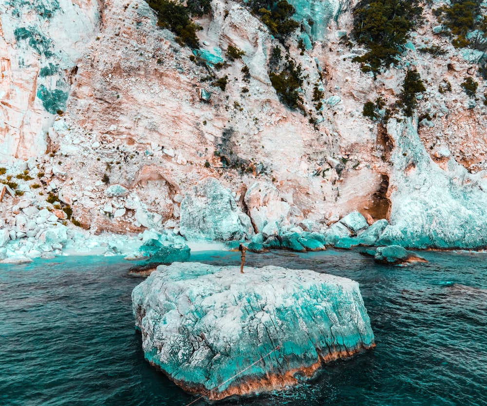 person in red shirt standing on rock formation in the middle of sea during daytime