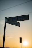 A street sign at an intersection points towards 'Hillston CCT' and 'Village CCT'. The background displays a clear sky with power lines, silhouetted against the warm tones of a setting or rising sun.