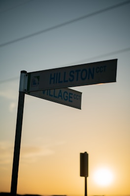 A street sign at an intersection points towards 'Hillston CCT' and 'Village CCT'. The background displays a clear sky with power lines, silhouetted against the warm tones of a setting or rising sun.