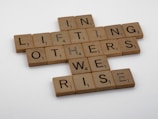 Scrabble tiles arranged on a white surface form the phrase 'In lifting others we rise'. Each tile has a wooden texture and displays both a letter and a corresponding point value.