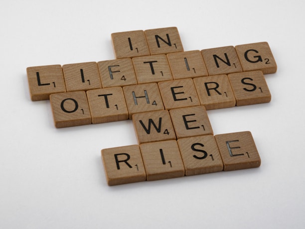 Scrabble tiles arranged on a white surface form the phrase 'In lifting others we rise'. Each tile has a wooden texture and displays both a letter and a corresponding point value.
