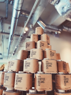 A display of stacked cylindrical containers labeled 'Coffee Beans' in a variety of numbers. The containers are made of cardboard with black text and are arranged to form a pyramid shape. In the background, metal ceiling pipes are visible, suggesting an industrial setting.