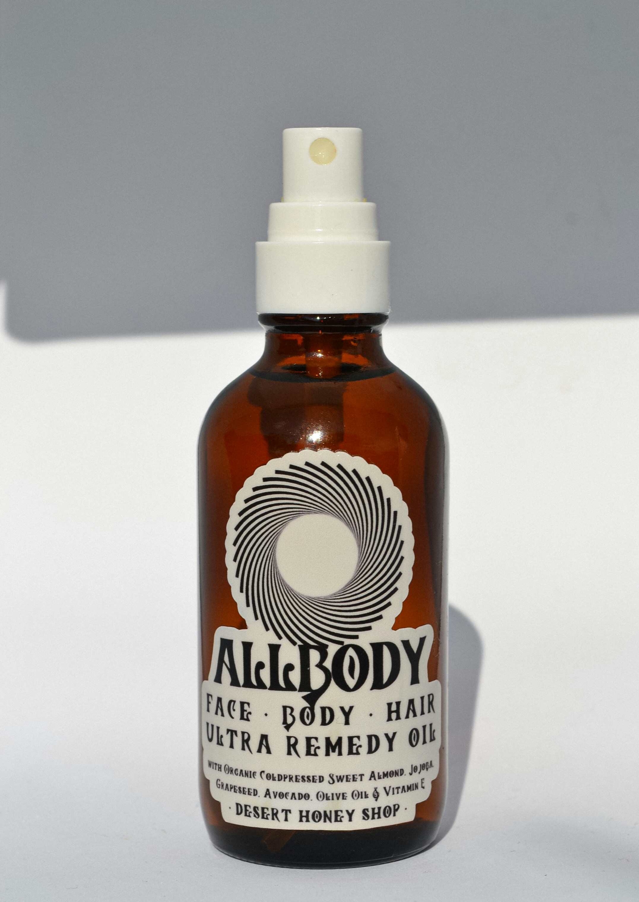 A brown glass bottle with a white spray nozzle containing a product labeled 'Allbody' for face, body, and hair. The label mentions ingredients such as organic cold-pressed sweet almond, jojoba, grapeseed, avocado, olive oil, and vitamin E. The bottle casts a soft shadow against a light background.