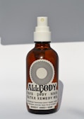 A brown glass bottle with a white spray nozzle containing a product labeled 'Allbody' for face, body, and hair. The label mentions ingredients such as organic cold-pressed sweet almond, jojoba, grapeseed, avocado, olive oil, and vitamin E. The bottle casts a soft shadow against a light background.