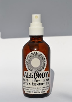A brown glass bottle with a white spray nozzle containing a product labeled 'Allbody' for face, body, and hair. The label mentions ingredients such as organic cold-pressed sweet almond, jojoba, grapeseed, avocado, olive oil, and vitamin E. The bottle casts a soft shadow against a light background.