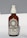 A brown glass bottle with a white spray nozzle containing a product labeled 'Allbody' for face, body, and hair. The label mentions ingredients such as organic cold-pressed sweet almond, jojoba, grapeseed, avocado, olive oil, and vitamin E. The bottle casts a soft shadow against a light background.