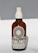 A brown glass bottle with a white spray nozzle containing a product labeled 'Allbody' for face, body, and hair. The label mentions ingredients such as organic cold-pressed sweet almond, jojoba, grapeseed, avocado, olive oil, and vitamin E. The bottle casts a soft shadow against a light background.