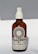 A brown glass bottle with a white spray nozzle containing a product labeled 'Allbody' for face, body, and hair. The label mentions ingredients such as organic cold-pressed sweet almond, jojoba, grapeseed, avocado, olive oil, and vitamin E. The bottle casts a soft shadow against a light background.