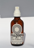 A brown glass bottle with a white spray nozzle containing a product labeled 'Allbody' for face, body, and hair. The label mentions ingredients such as organic cold-pressed sweet almond, jojoba, grapeseed, avocado, olive oil, and vitamin E. The bottle casts a soft shadow against a light background.