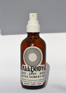 A brown glass bottle with a white spray nozzle containing a product labeled 'Allbody' for face, body, and hair. The label mentions ingredients such as organic cold-pressed sweet almond, jojoba, grapeseed, avocado, olive oil, and vitamin E. The bottle casts a soft shadow against a light background.