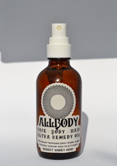 A brown glass bottle with a white spray nozzle containing a product labeled 'Allbody' for face, body, and hair. The label mentions ingredients such as organic cold-pressed sweet almond, jojoba, grapeseed, avocado, olive oil, and vitamin E. The bottle casts a soft shadow against a light background.
