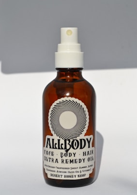 A brown glass bottle with a white spray nozzle containing a product labeled 'Allbody' for face, body, and hair. The label mentions ingredients such as organic cold-pressed sweet almond, jojoba, grapeseed, avocado, olive oil, and vitamin E. The bottle casts a soft shadow against a light background.