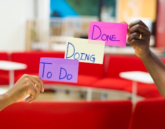 Three hands hold up colorful sticky notes labeled 'To Do', 'Doing', and 'Done' against a blurred background of red seating and a modern office environment.