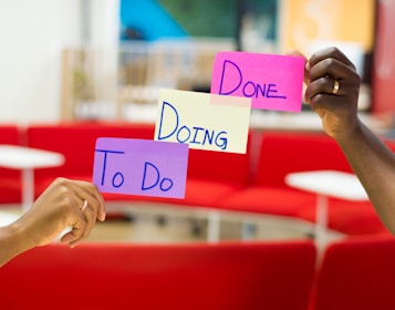 Three hands hold up colorful sticky notes labeled 'To Do', 'Doing', and 'Done' against a blurred background of red seating and a modern office environment.
