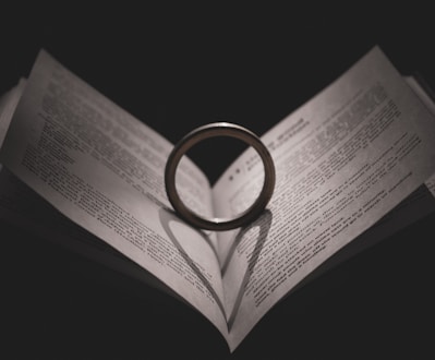 A book is open with its pages slightly creased, forming a heart shape. At the center, a ring stands upright, casting a heart-shaped shadow onto the pages below. The lighting is dark, creating a dramatic and romantic ambiance.