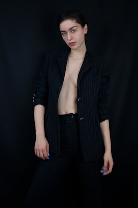 A person stands against a dark background wearing a pinstripe suit jacket that is open to reveal part of their torso. The person has a neutral expression and their nails are painted blue.
