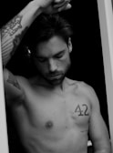 A black and white portrait of a shirtless man with tattoos. He appears deep in thought with closed eyes and his right arm raised, resting on something out of frame. The tattoos include a stylized design on his forearm and the number 42 on his chest.