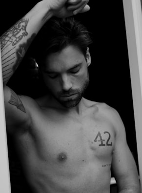 A black and white portrait of a shirtless man with tattoos. He appears deep in thought with closed eyes and his right arm raised, resting on something out of frame. The tattoos include a stylized design on his forearm and the number 42 on his chest.