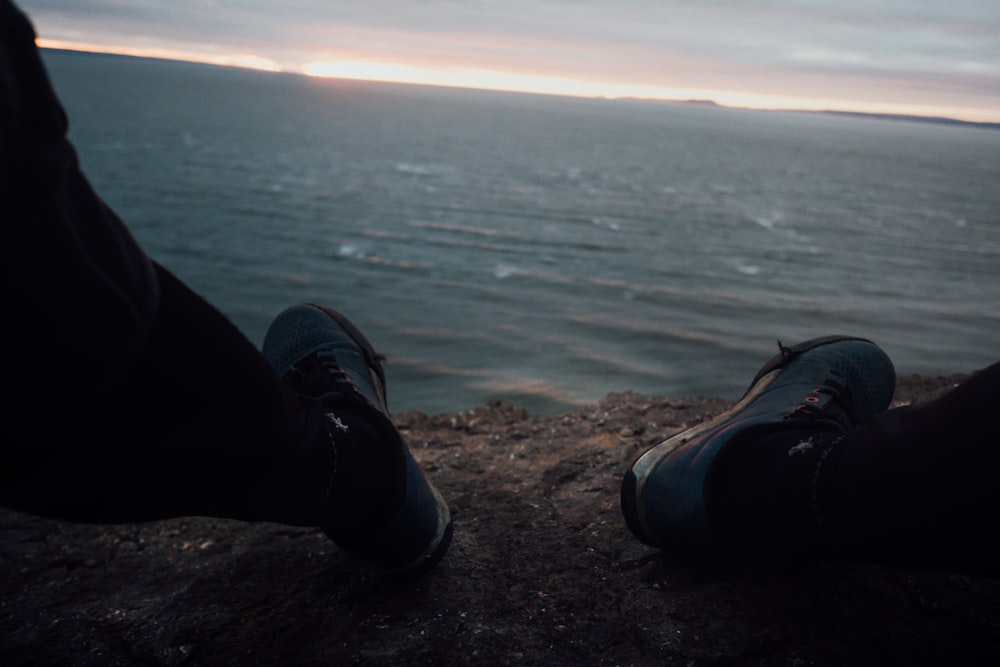 person in black pants and black and white sneakers sitting on rock near body of water