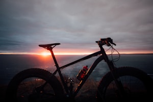 A silhouette of a bicycle with wide tires stands against a dramatic sunset over an ocean horizon. The sky is filled with clouds that are tinged with orange and purple hues, creating a striking contrast with the dark outline of the bicycle. The overall scene suggests a tranquil and adventurous setting.
