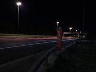 A nighttime highway scene with bright streetlights illuminating the road. An SOS emergency call box stands prominently on the roadside. Light trails from passing vehicles create streaks of color on the asphalt.