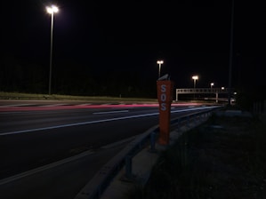 A nighttime highway scene with bright streetlights illuminating the road. An SOS emergency call box stands prominently on the roadside. Light trails from passing vehicles create streaks of color on the asphalt.