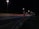A nighttime highway scene with bright streetlights illuminating the road. An SOS emergency call box stands prominently on the roadside. Light trails from passing vehicles create streaks of color on the asphalt.