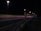 A nighttime highway scene with bright streetlights illuminating the road. An SOS emergency call box stands prominently on the roadside. Light trails from passing vehicles create streaks of color on the asphalt.