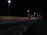 A nighttime highway scene with bright streetlights illuminating the road. An SOS emergency call box stands prominently on the roadside. Light trails from passing vehicles create streaks of color on the asphalt.